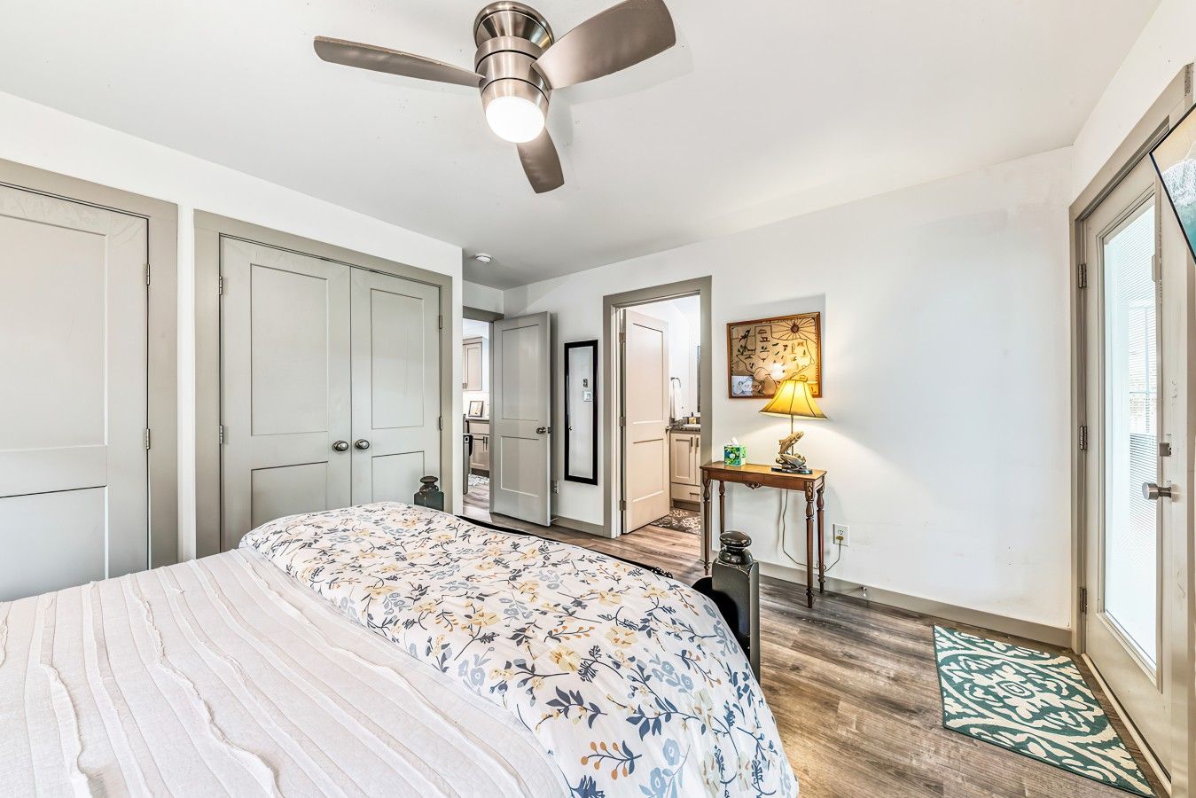 A bedroom with a bed, a ceiling fan, a bedside table with a lamp, and a door leading to another room.