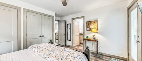 A bedroom with a bed, a ceiling fan, a bedside table with a lamp, and a door leading to another room.
