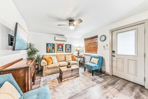 Uma sala de estar com sofá, poltronas, mesa de centro, televisão, ventoinha no teto, janela com persianas e uma porta.