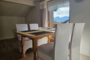 A dining area with a wooden table and white chairs, a window with a view of mountains, and a curtain with a checkered pattern. - Holiday apartment Breitenberg (Hopferau)