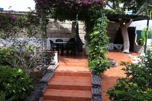 Een terras met een stenen trap naar een tafel en stoelen, omgeven door groen en een pergola.