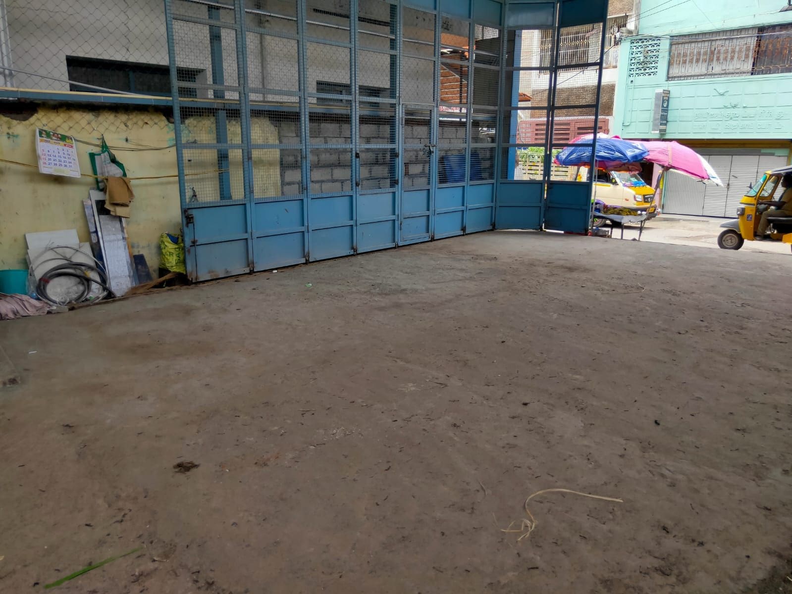 Un espacio al aire libre rodeado por una cerca, con suelo de tierra, un auto-rickshaw amarillo y un edificio azul al fondo.