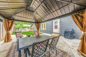 A covered patio with a table and chairs, a barbecue grill, and a house in the background.