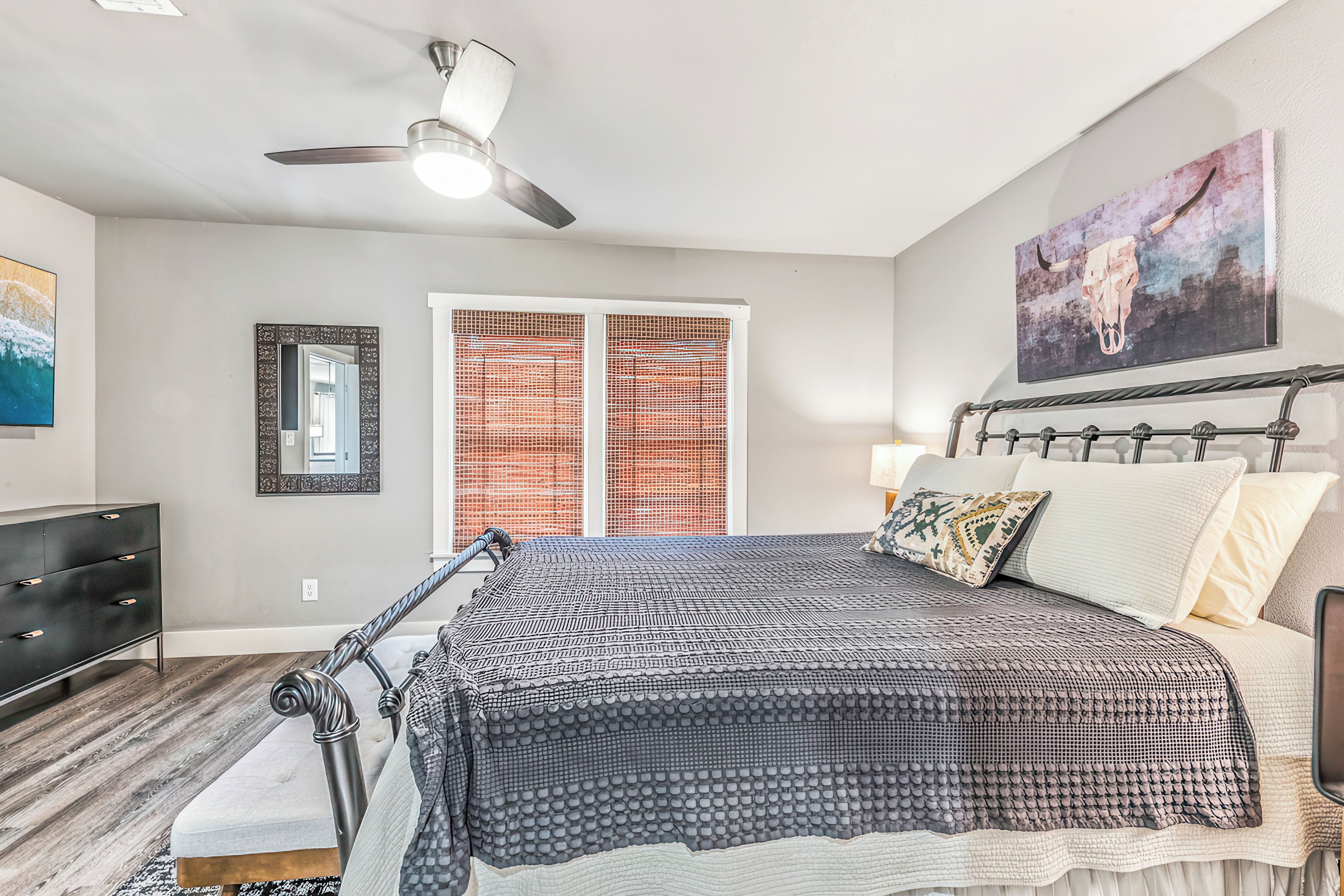A bedroom with a bed, a ceiling fan, a window with blinds, a dresser, and a painting on the wall.