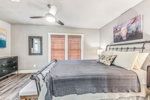 A bedroom with a bed, a ceiling fan, a window with blinds, a dresser, and a painting on the wall.