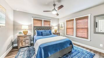 Une chambre à coucher comprenant un lit, des tables de chevet, un ventilateur de plafond et une fenêtre avec des stores.