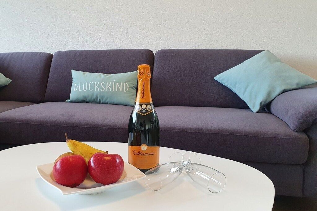A bottle of champagne, two apples, and a glass on a white table with a purple sofa in the background.