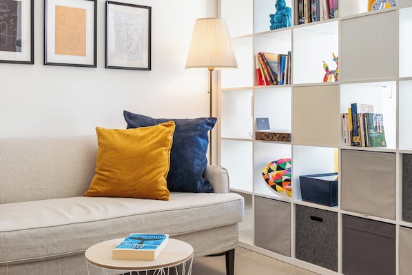 A living room with a sofa, cushions, a small round table with a book, a floor lamp, and a bookshelf with various items. - Modern Elegant UNIC Studio Suite (Egkomi Lefkosias)