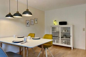 Un coin repas avec une table blanche, des chaises jaunes, des suspensions noires et un buffet blanc doté de portes en verre.