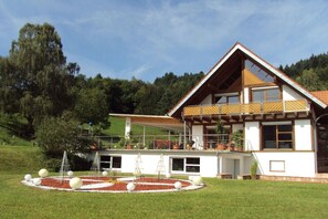 A two-story house with a balcony, a circular garden with spherical decorations, and a forested hillside in the background. - Exclusive holiday home with pool (Berghaupten)