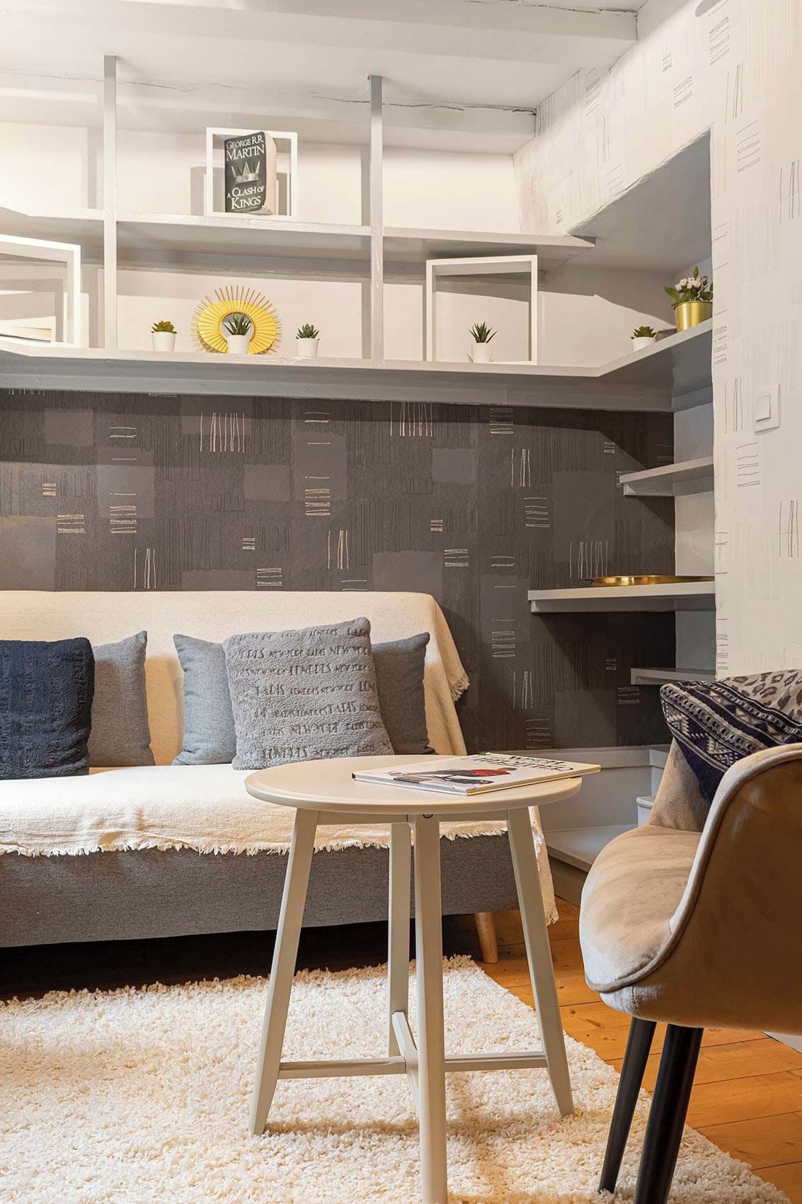 A modern living room with a sofa, a round table, and a chair. There are shelves with decorative items and a wall with a geometric pattern.