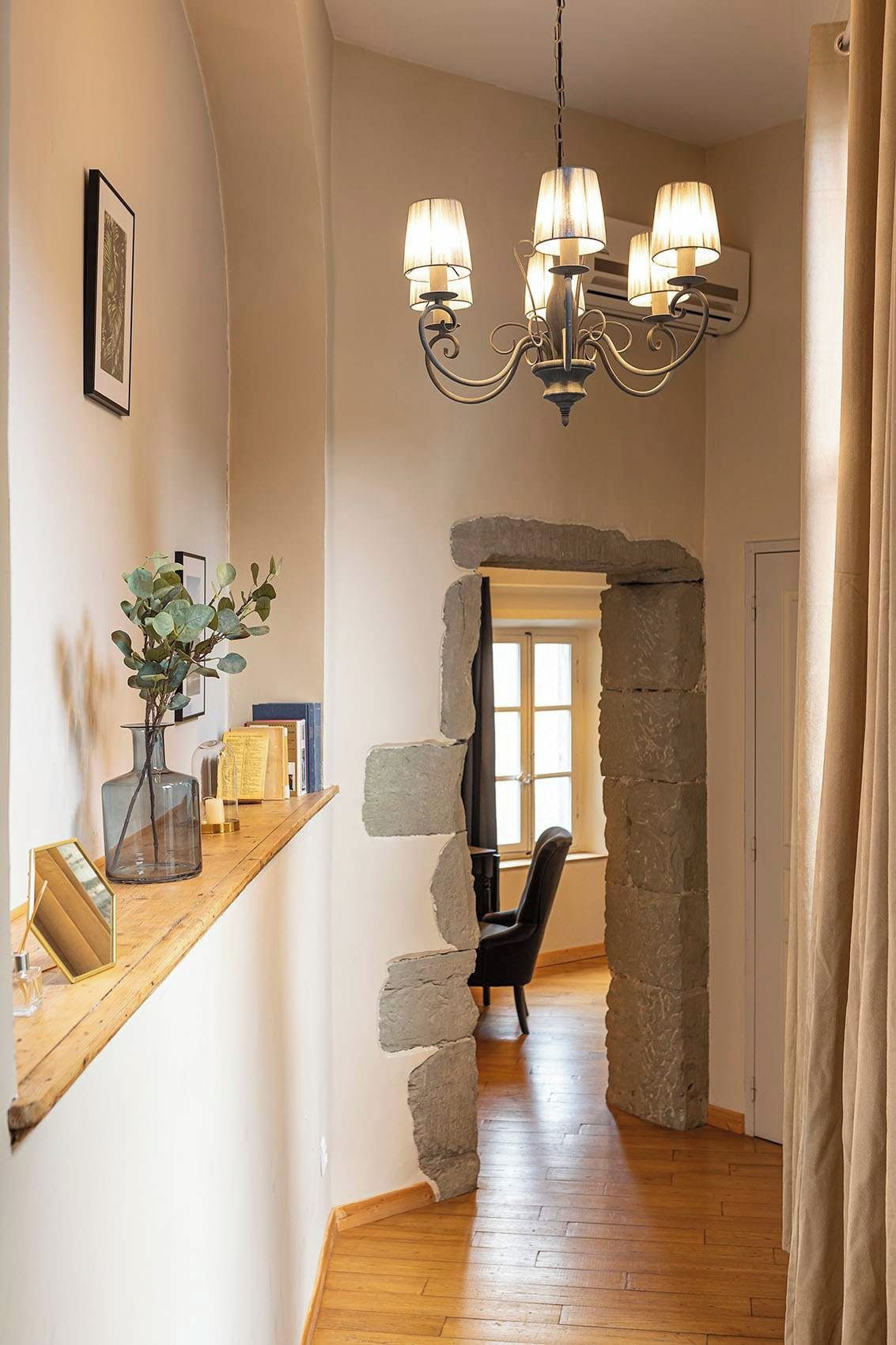 Um corredor com parede de pedra, prateleira de madeira com livros e um vaso, uma cadeira preta e um lustre.