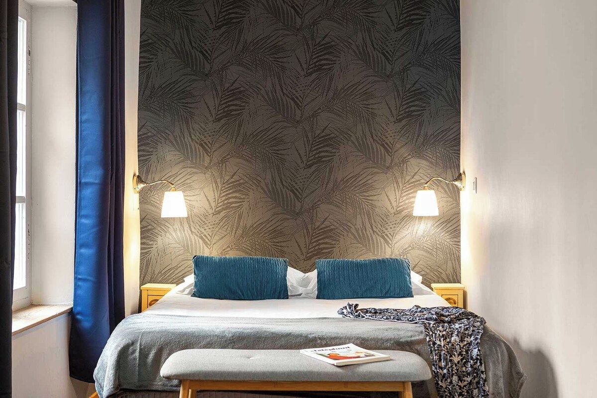 Un dormitorio con cama, dos lámparas de noche, una ventana con cortinas y un papel tapiz estampado.