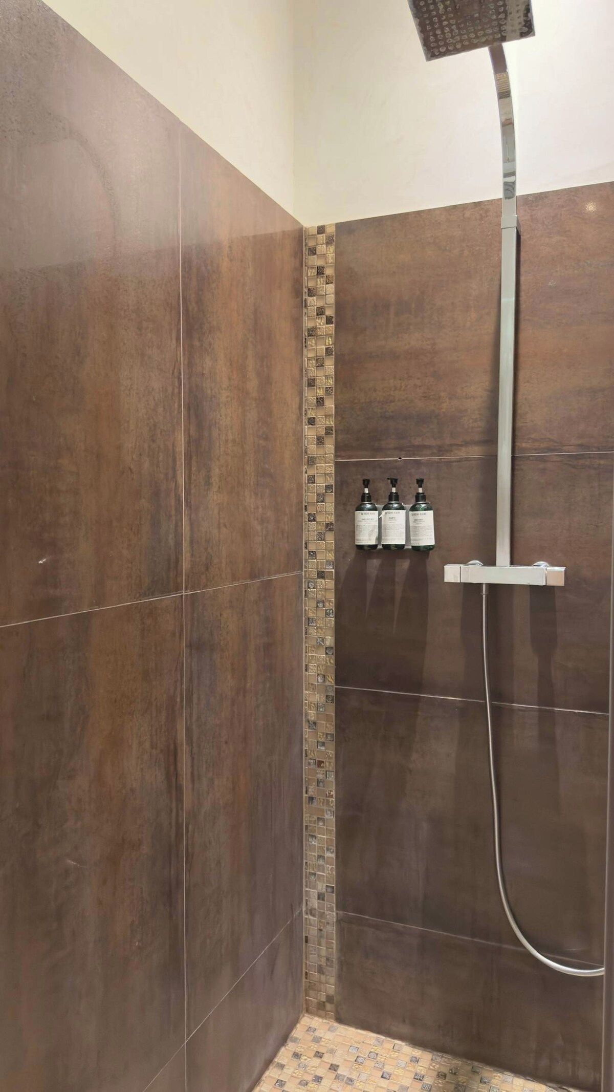 Een douchegedeelte met een betegelde muur, een douchekop en drie flessen shampoo.