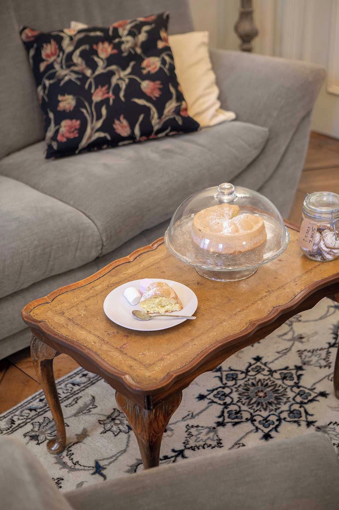 Mesa de jantar com bolo e prato de comida, sofá com almofada floral e tapete estampado.