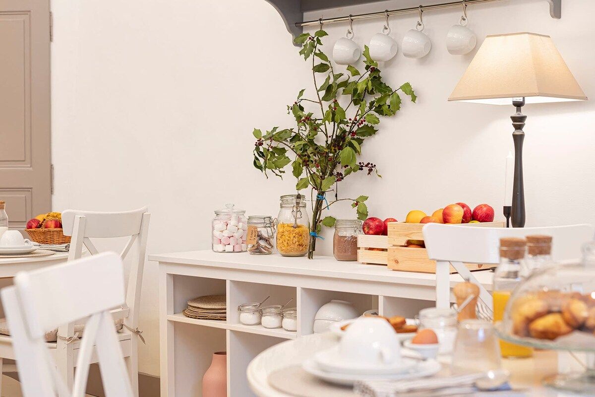 A dining area with white furniture, a table with fruits, a vase with greenery, and a lamp.
