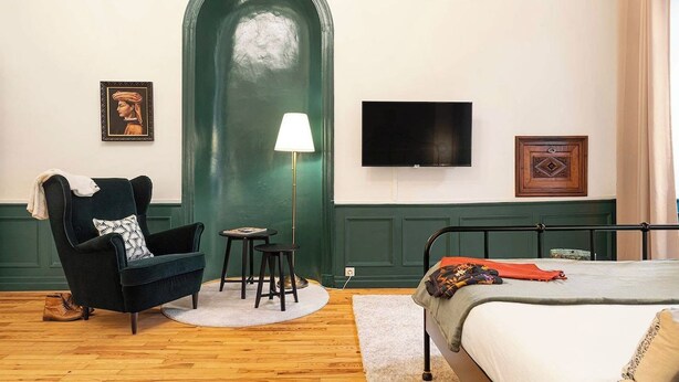 Ein Zimmer mit einem Bett, einem Sessel, einer Lampe, einem Fernseher und einer grünen Tür.