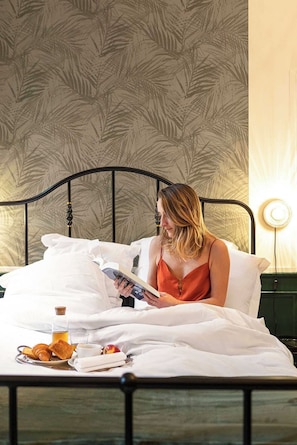 A person sitting on a bed with a book, a bottle, and food items on the bedside table. - Chambre Verveine Chambéry (Chambéry)
