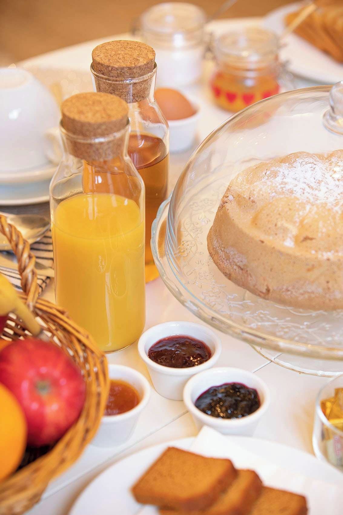 Mesa de café da manhã com um suporte de vidro para bolo, uma garrafa de suco de laranja e potinhos de geleia.