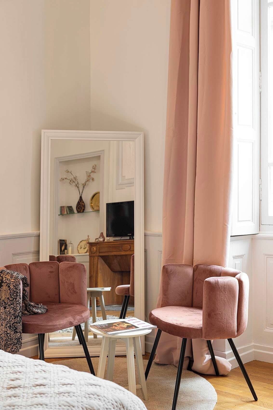 Une pièce comprenant deux fauteuils roses, une petite table ronde, un fauteuil blanc avec un coussin à motifs, et une porte blanche ornée d’un miroir et d’objets décoratifs.