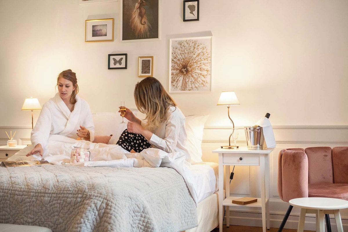 Habitación de hotel con una cama, mesitas de noche, un sillón rosa y obras de arte en las paredes.