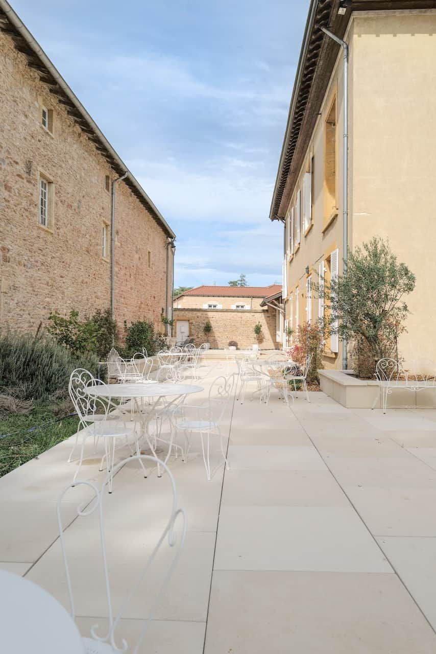 An outdoor patio with white metal chairs and tables, surrounded by stone buildings.