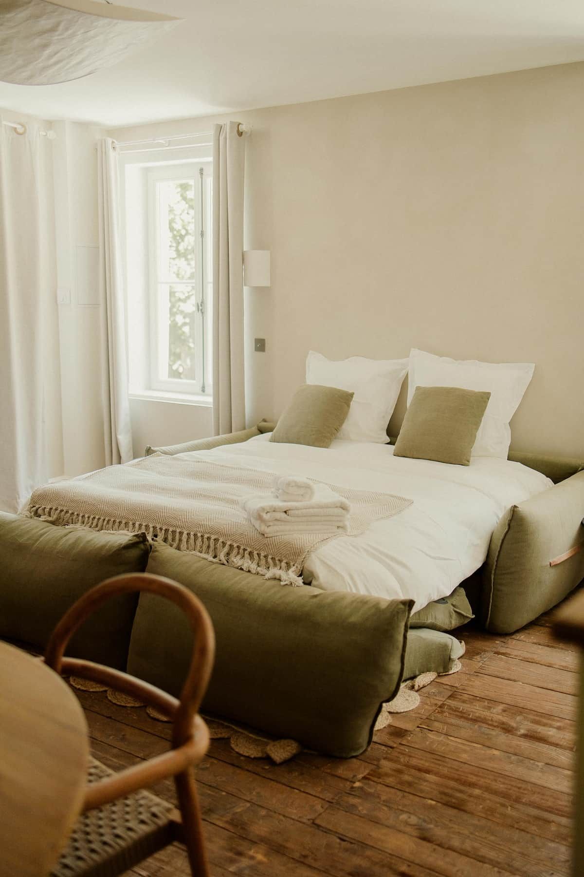 Een slaapkamer met een bed, kussens, een deken, een stoel en een raam.