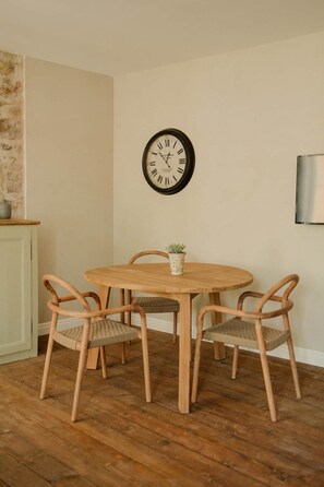 Een eetgedeelte met een ronde houten tafel, vier rieten stoelen, een wandklok en een kleine plant in een pot.