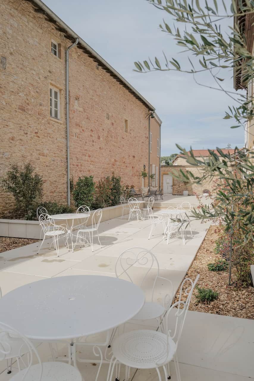 Eine Außenpatio-Terrasse mit weißen Metalltischen und -stühlen, ein Backsteinhaus im Hintergrund und etwas Grün.