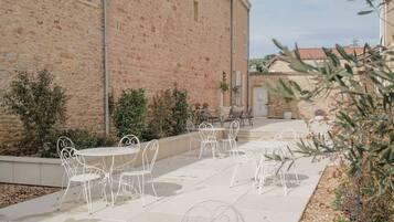 Eine Außenpatio-Terrasse mit weißen Metalltischen und -stühlen, ein Backsteinhaus im Hintergrund und etwas Grün.