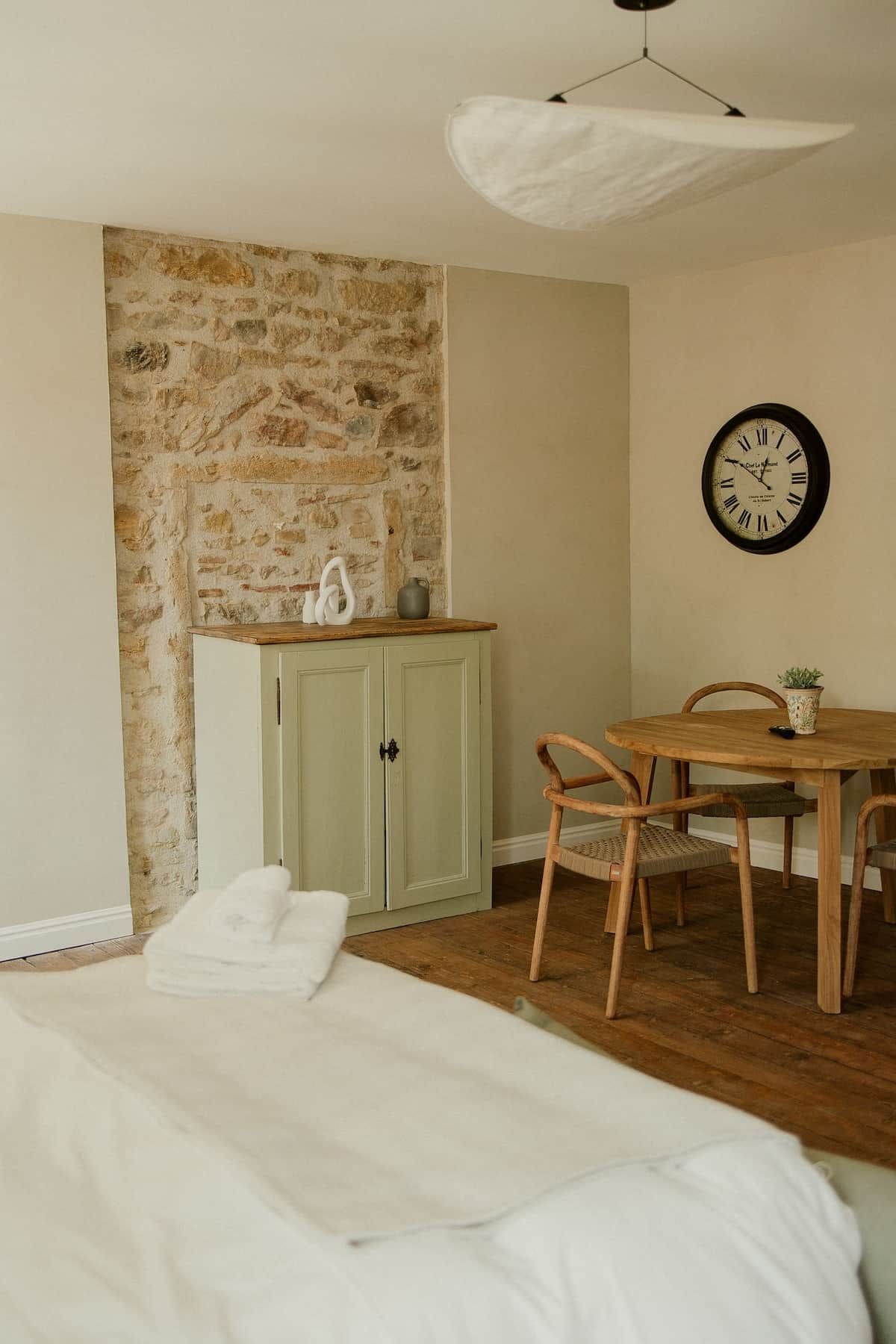 A room with a stone wall, a wooden table with chairs, a bed with white bedding, and a wall clock.