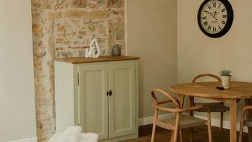 A room with a stone wall, a wooden table with chairs, a bed with white bedding, and a wall clock.