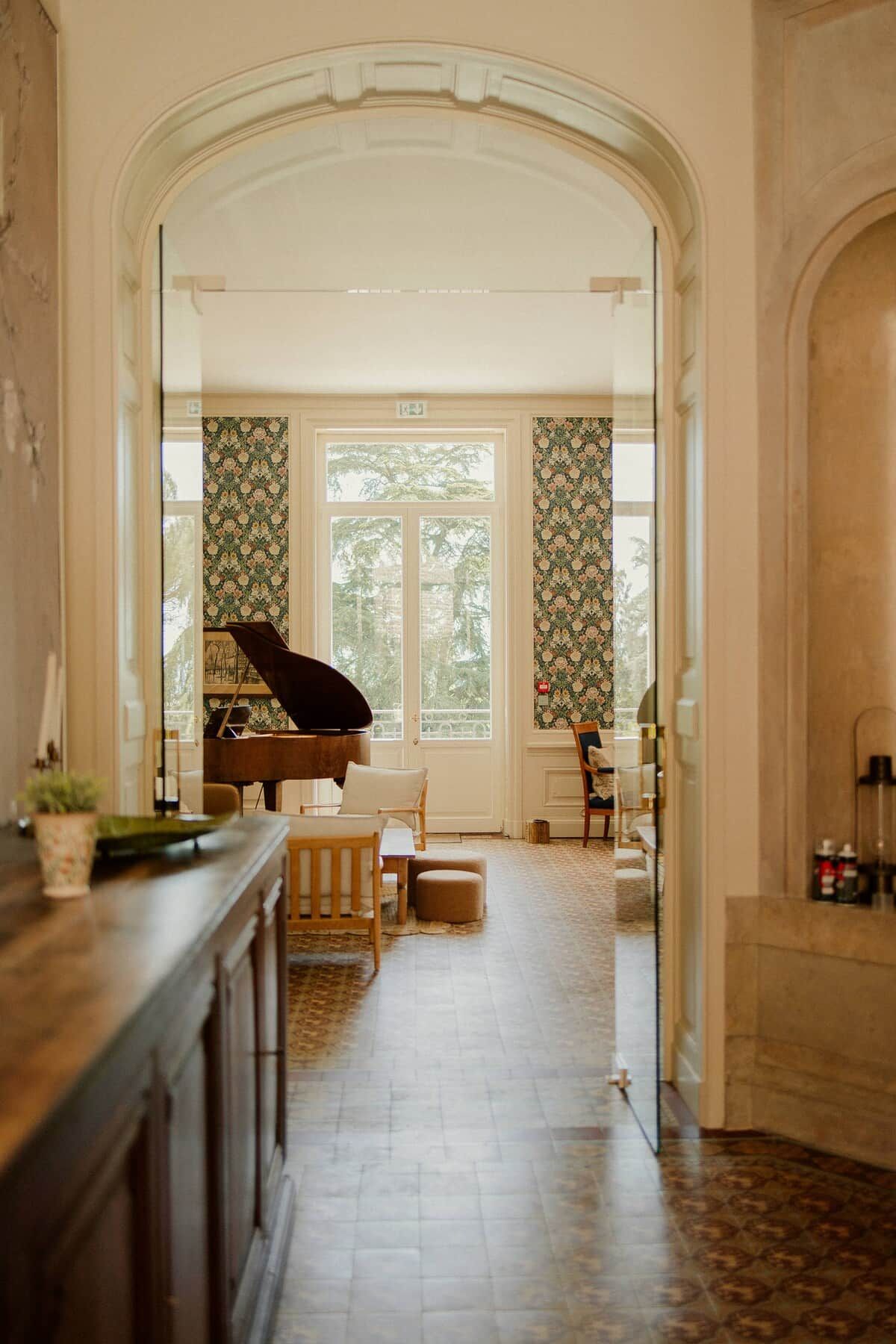 Een vleugelpiano in een ruime kamer met een groot raam, een open haard en behangpapier met een patroon.