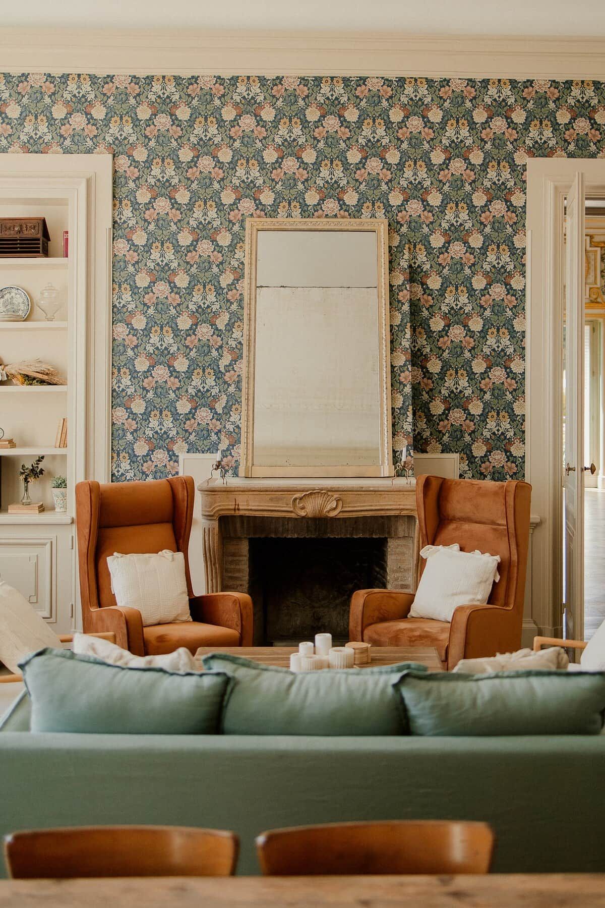 Une pièce avec une cheminée, deux fauteuils, un canapé vert et un papier peint à motifs.
