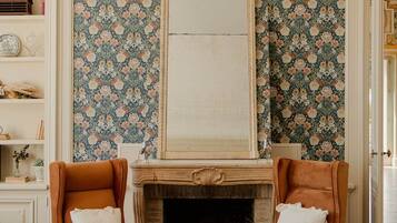 Une pièce avec une cheminée, deux fauteuils, un canapé vert et un papier peint à motifs.