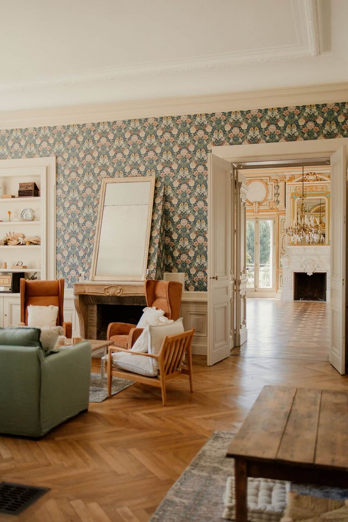 A spacious living room with a fireplace, wooden flooring, and a floral wallpaper design.
