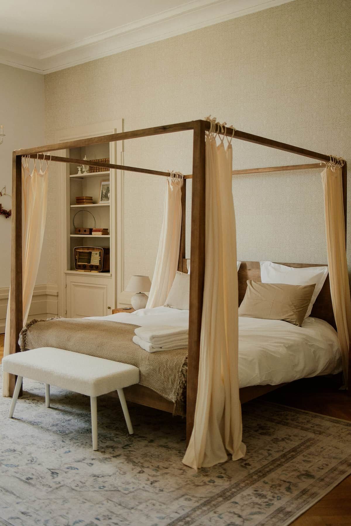 Un lit à baldaquin à quatre poteaux, agrémenté d’une table de chevet et d’un banc, d’une petite table de nuit et d’une petite étagère où sont disposés des objets décoratifs.