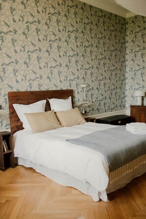 A neatly made bed with white and beige bedding, a wooden headboard, and a nightstand with a lamp.
