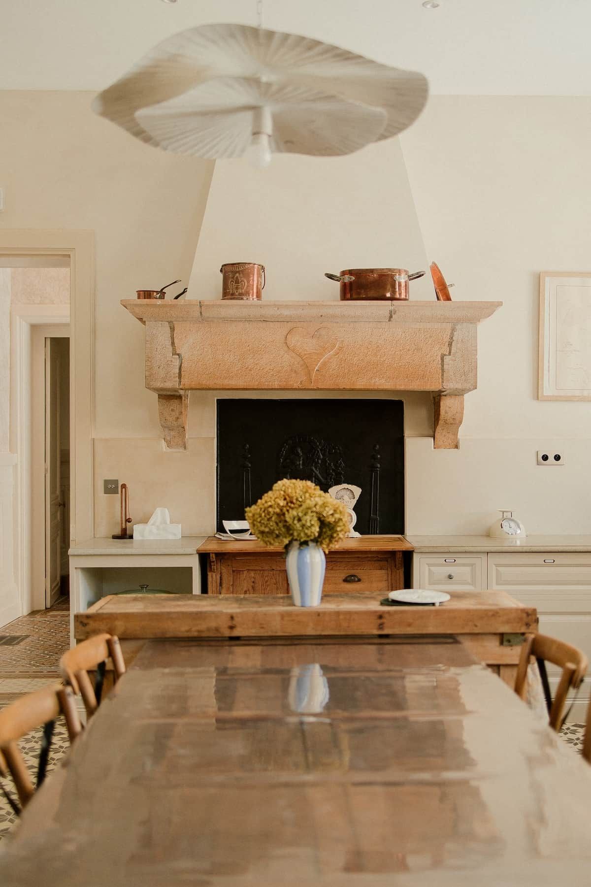 A dining area with a long wooden table, a vase with flowers, and a fireplace with copper pots above.