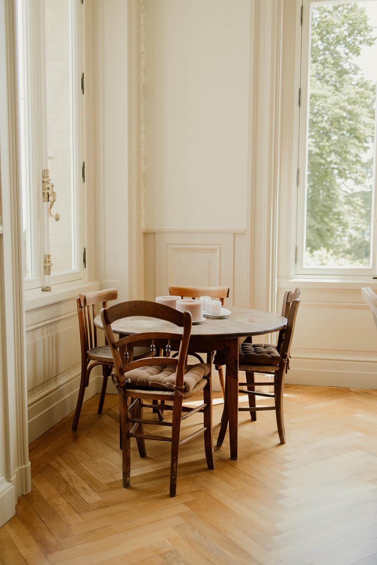Een eetkamer met een houten tafel en stoelen, een raam met uitzicht op bomen, en een deur met een klink van messing.