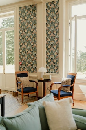 Ein Zimmer mit zwei Holzstühlen, einem kleinen Tisch mit Lampe und einem grünen Sofa.