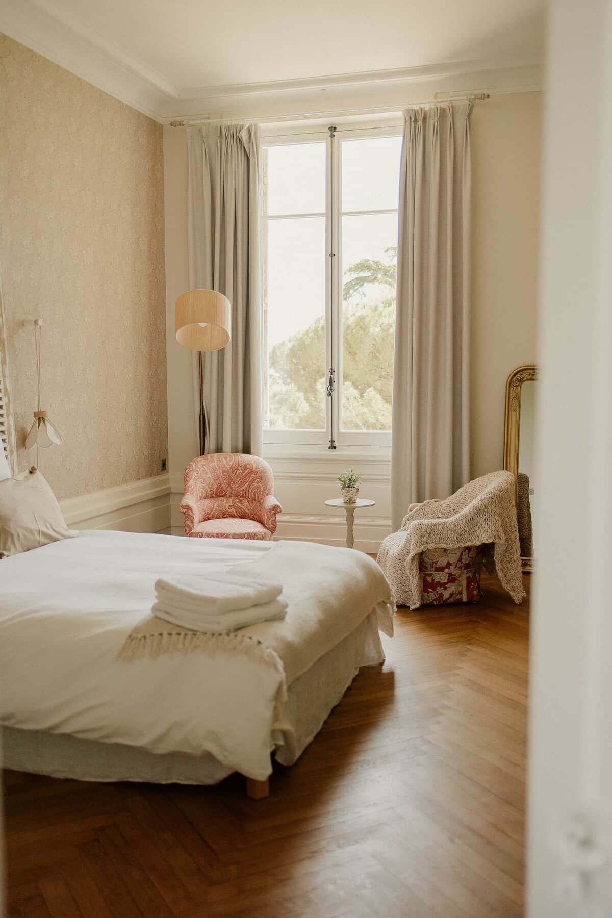 A bedroom with a large bed, a chair, a lamp, and a window with curtains.