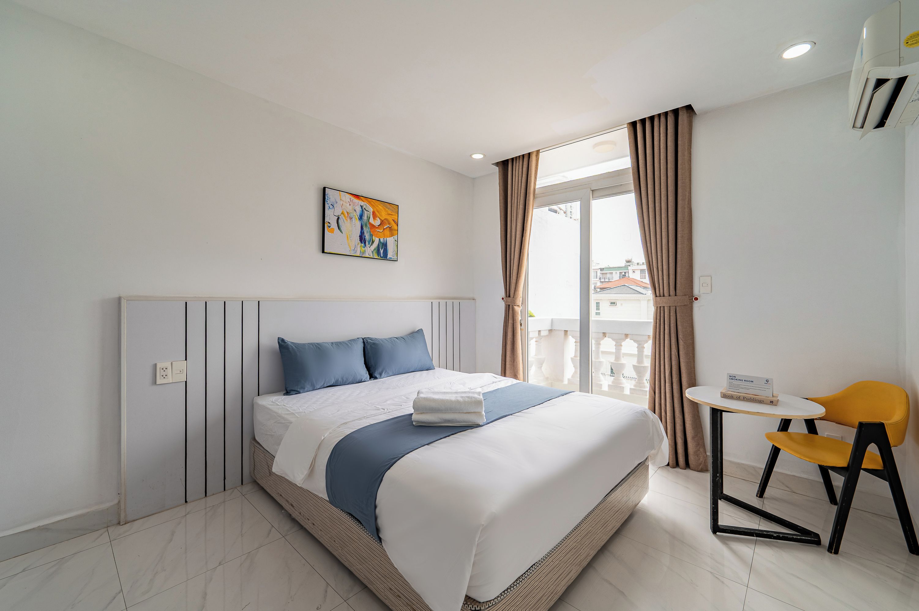 Habitación de hotel con cama, un cuadro, una ventana con cortinas, una mesita con una silla y un cartel que indica que no se permite fumar.