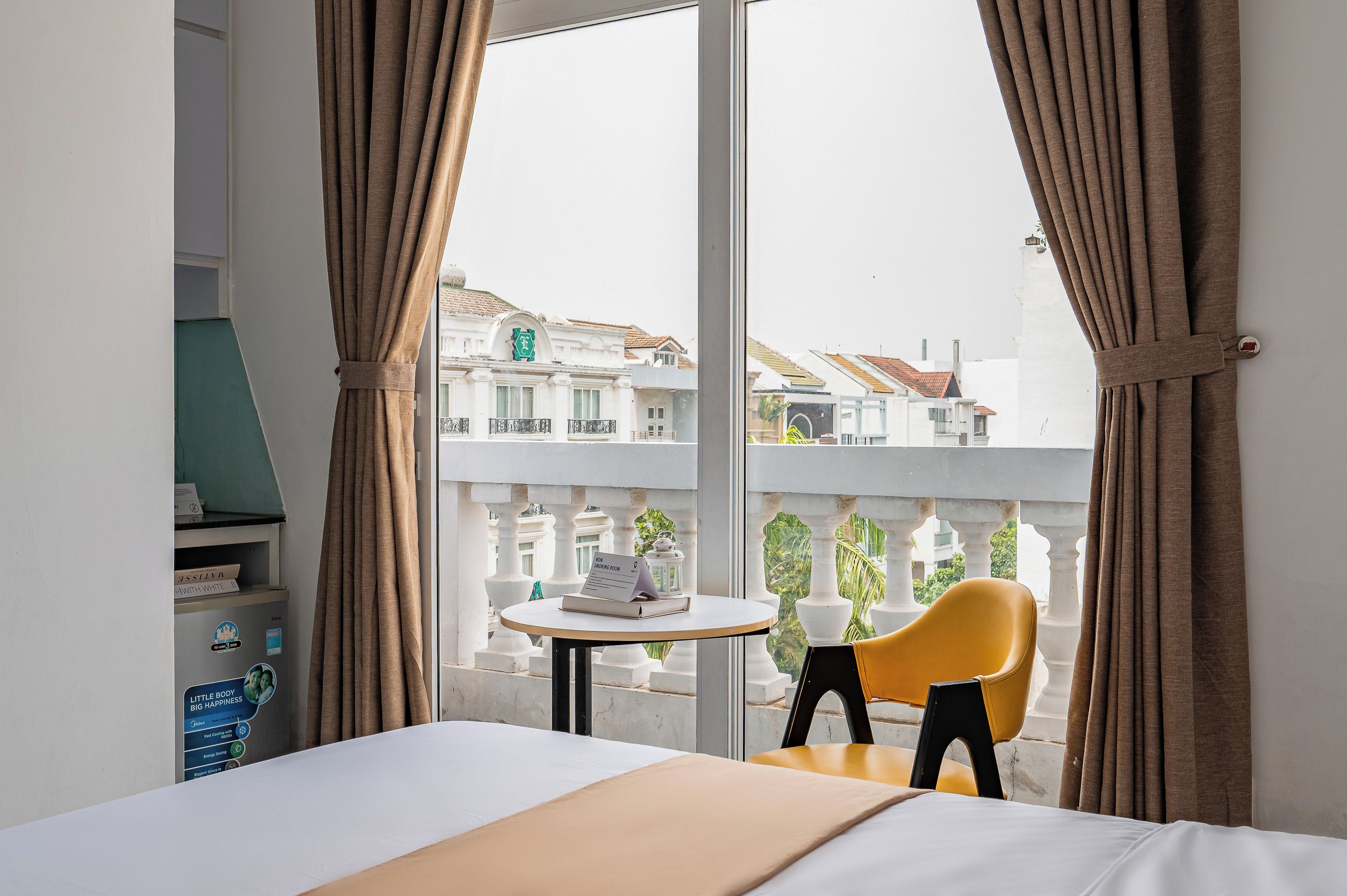 A hotel room with a bed, a small table with a sign, a yellow chair, and a view of buildings outside.