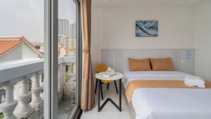 A hotel room with a bed, a small table, a chair, a painting, and a balcony with a view of buildings. - Cozrum Homes - The Ivory House (Tan Hung)