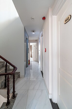 A hallway with a marble floor, a wooden staircase, and a fire alarm on the wall. - Cozrum Homes - The Ivory House (Tan Hung)