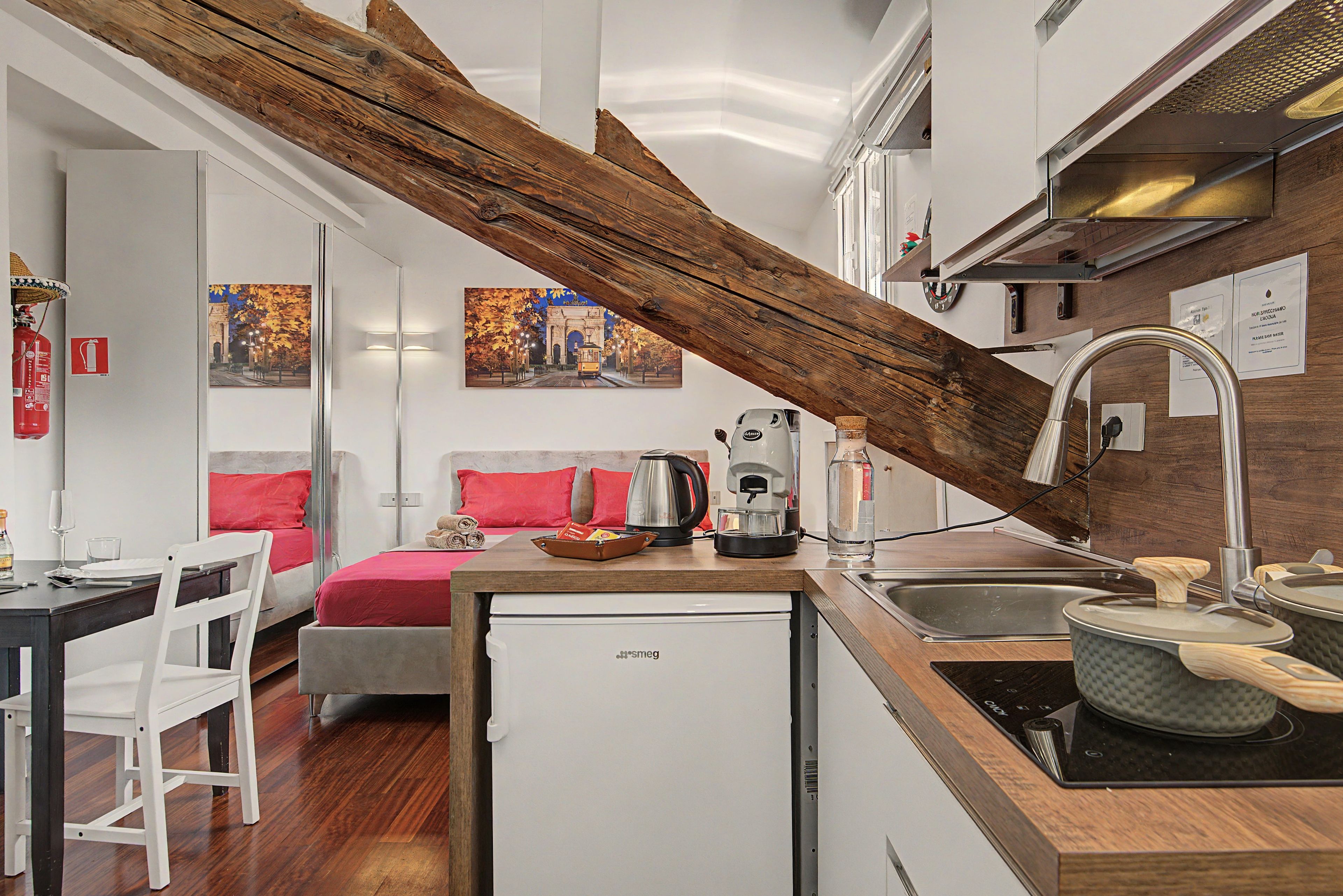 A compact hotel room with a kitchenette, a bed with red bedding, a small dining area, and a wooden beam ceiling.