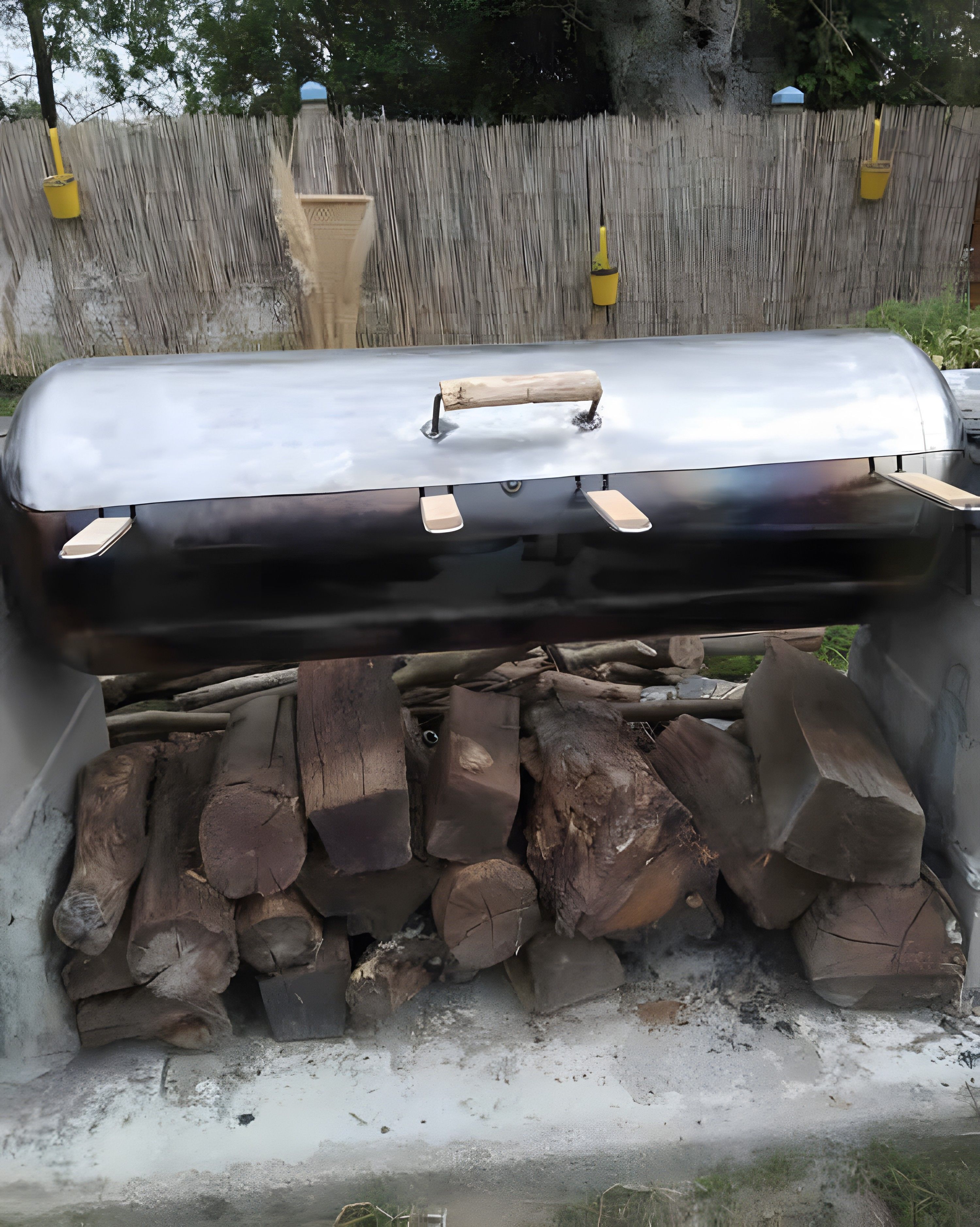 Una parrilla para barbacoa con un asa, ubicada al aire libre con una valla de madera y luces amarillas colgantes al fondo.