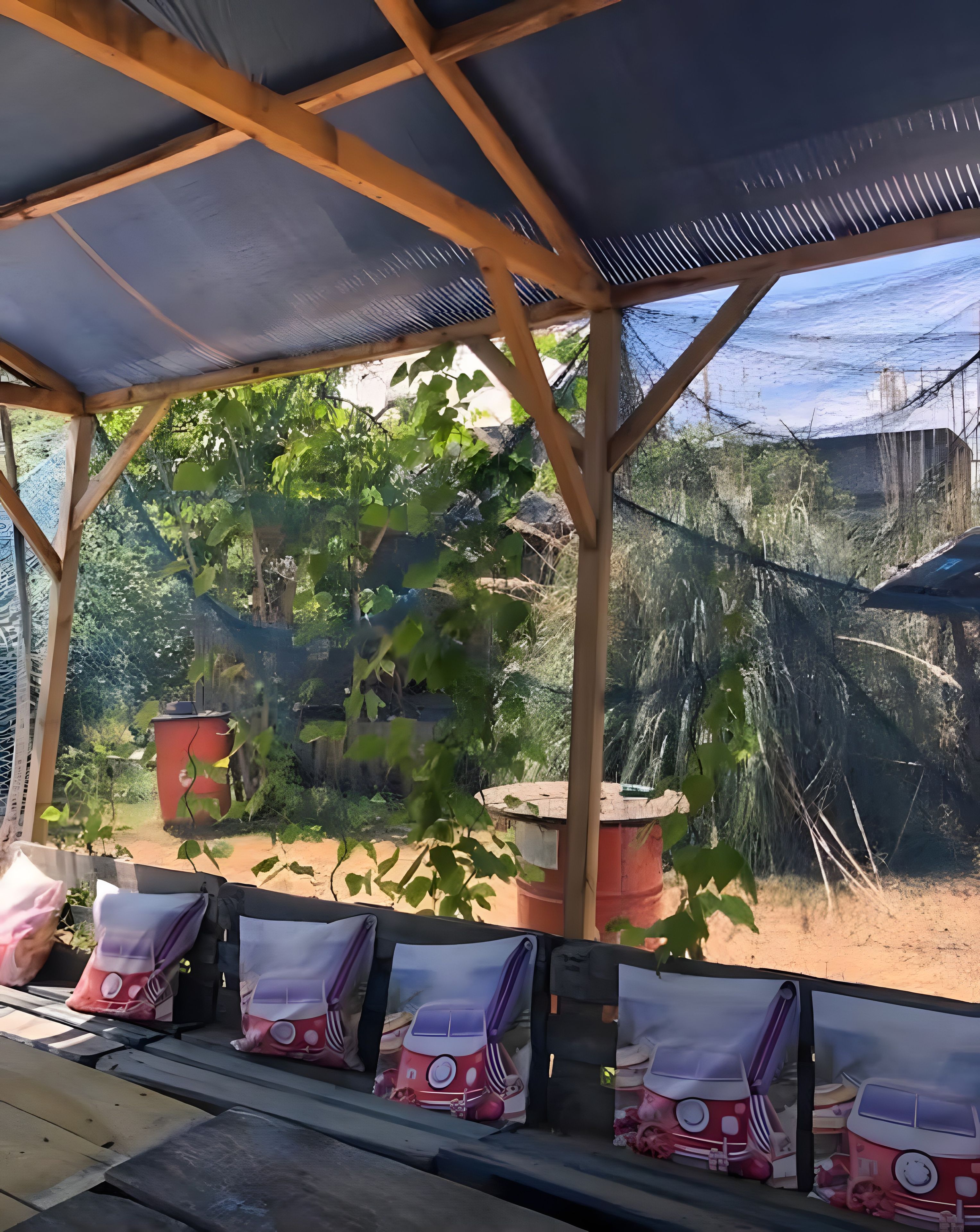 A covered outdoor seating area with wooden beams and a blue roof, surrounded by greenery and a view of a natural landscape.