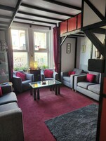 Una sala de estar amoblada con una mesa de centro de vidrio, dos sofás grises con cojines rojos, una alfombra roja y vistas a la calle a través de amplias ventanas.